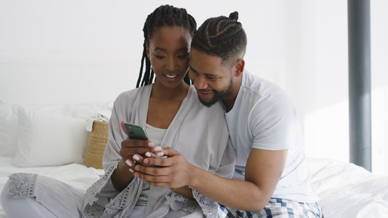 一对幸福的年轻夫妇坐在家里的床上，一起使用智能手机。恋爱中的男女，醒来后在卧室里拿着手机上网视频素材