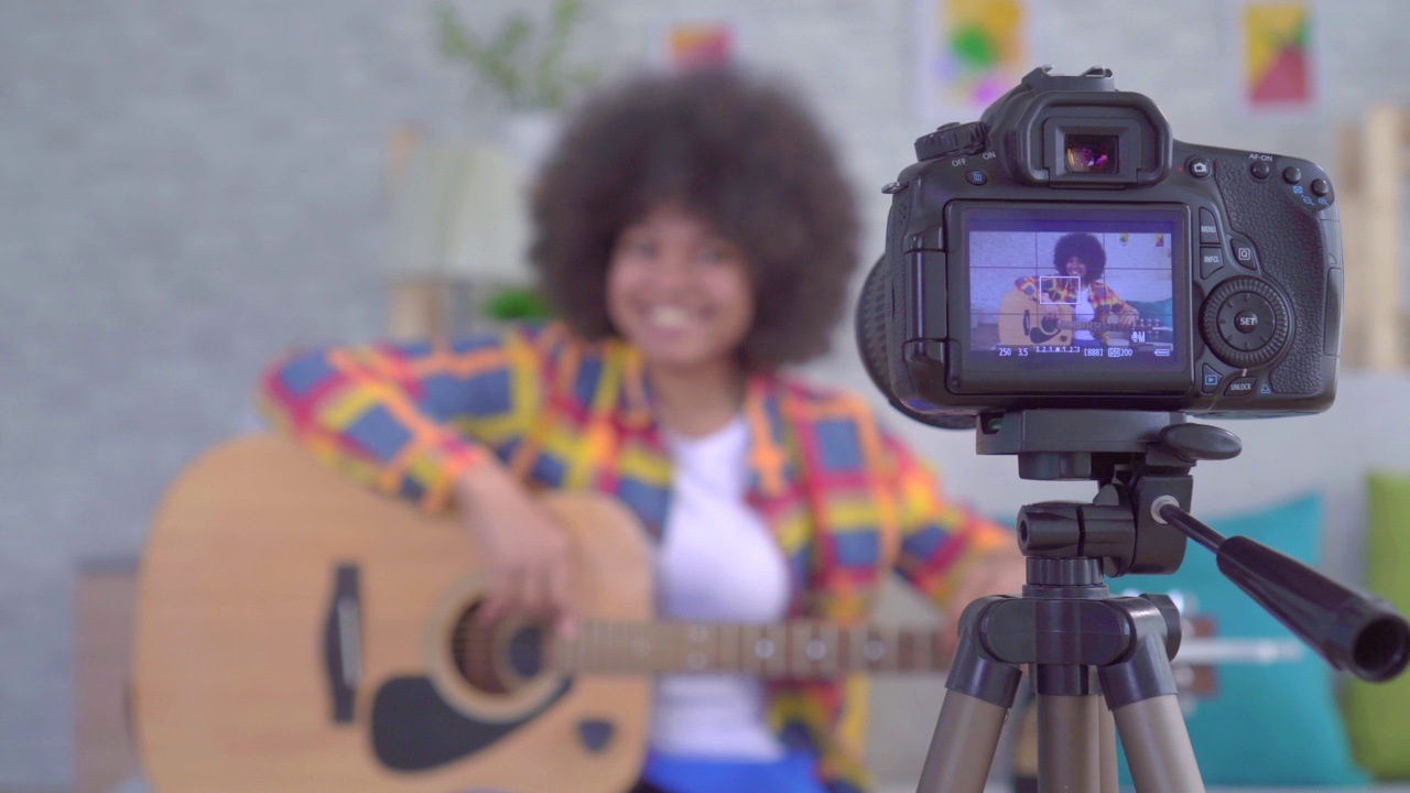 非洲裔美国女性博主，留着非洲式发型，拿着一把吉他，透过相机屏幕观看视频下载