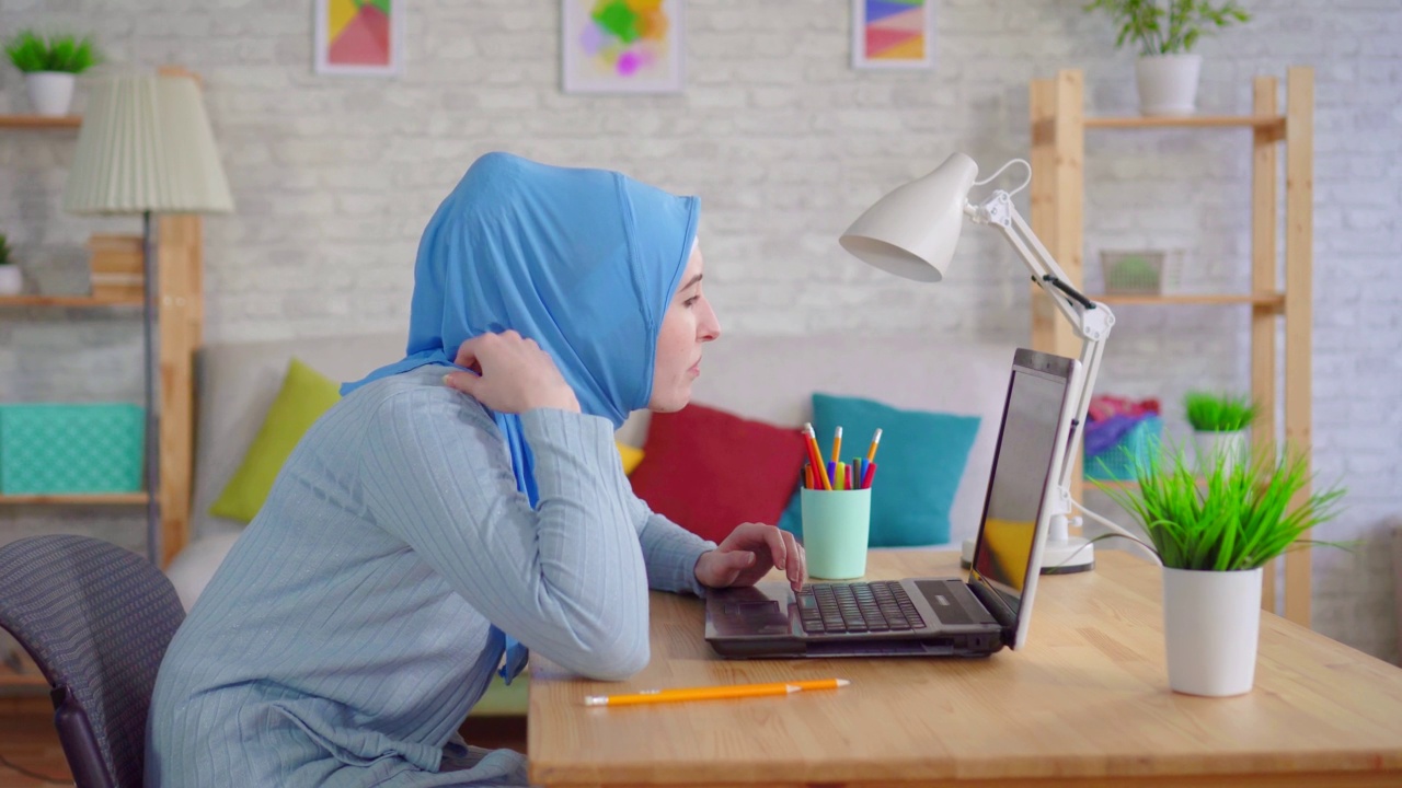 一名戴着传统头巾的年轻穆斯林妇女坐在笔记本电脑前无精打采，感到脖子和背部疼痛视频下载