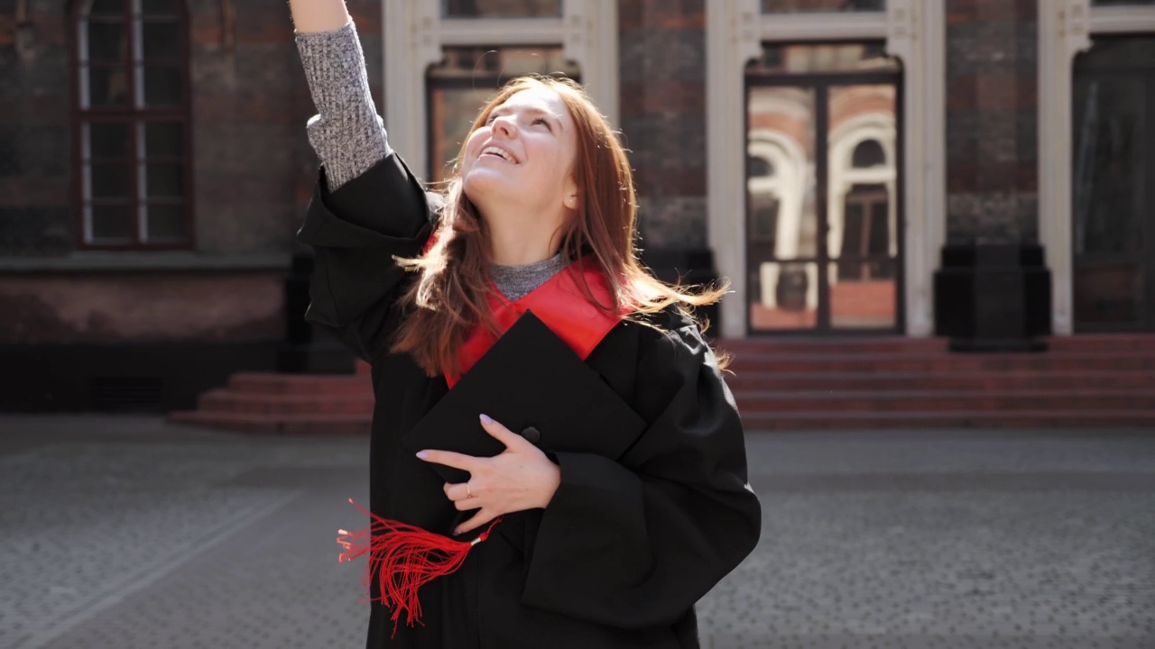 快乐美丽的姜黄女学生，披着斗篷，戴着硕士学位帽，手拿毕业证书，在高中毕业当天举起了手。视频素材
