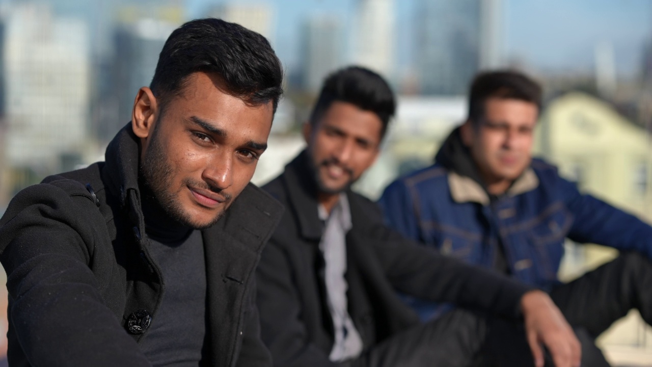 年轻的中东男子回头与模糊的朋友交谈看着相机。英俊自信的千禧一代男子的肖像在户外阳光下休息在屋顶上。视频下载