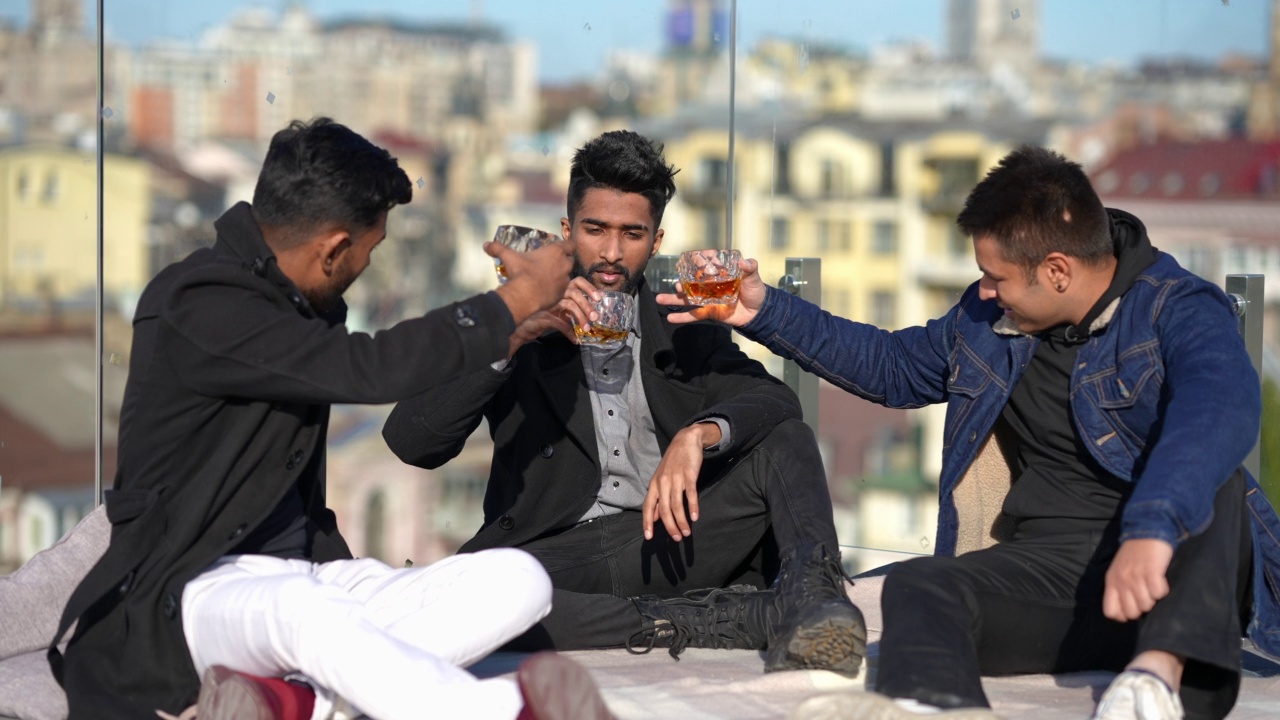 广角镜头失意的英俊年轻人和朋友坐在屋顶上碰杯喝威士忌。抑郁的中东千禧一代的肖像和朋友的支持。视频下载