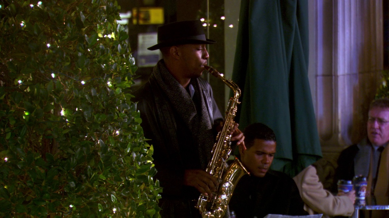 中等角度的街头爵士音乐家演奏萨克斯管前面的路边咖啡馆或餐馆。顾客，顾客坐在桌旁聊天，喝咖啡。用透明的灯装饰的盆栽或人造树。可以是假期还是圣诞节视频素材