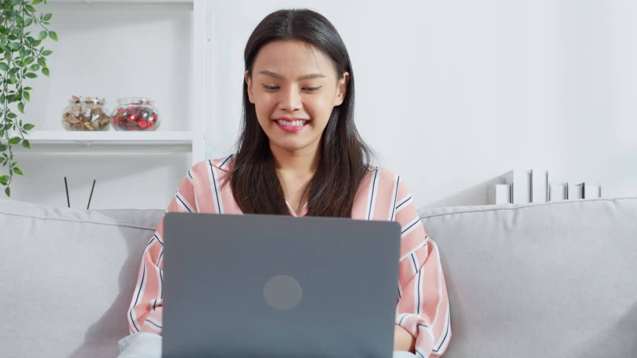 亚洲年轻女商人在电脑上打字，在家工作。漂亮迷人的自由职业女职工坐在沙发上，用笔记本电脑在客厅的幸福和微笑的房子视频素材