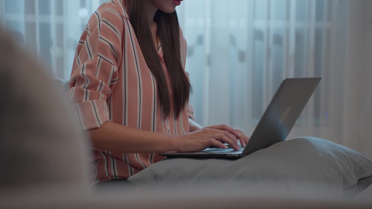 年轻的亚洲女商人在电脑上打字，在家工作。漂亮迷人的自由职业女职工坐在沙发上，用笔记本电脑在客厅的幸福和微笑的房子视频素材
