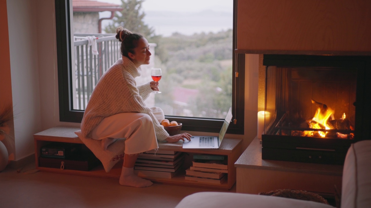 舒适的家。年轻女子在壁炉旁喝着红酒，用着笔记本电脑。视频下载