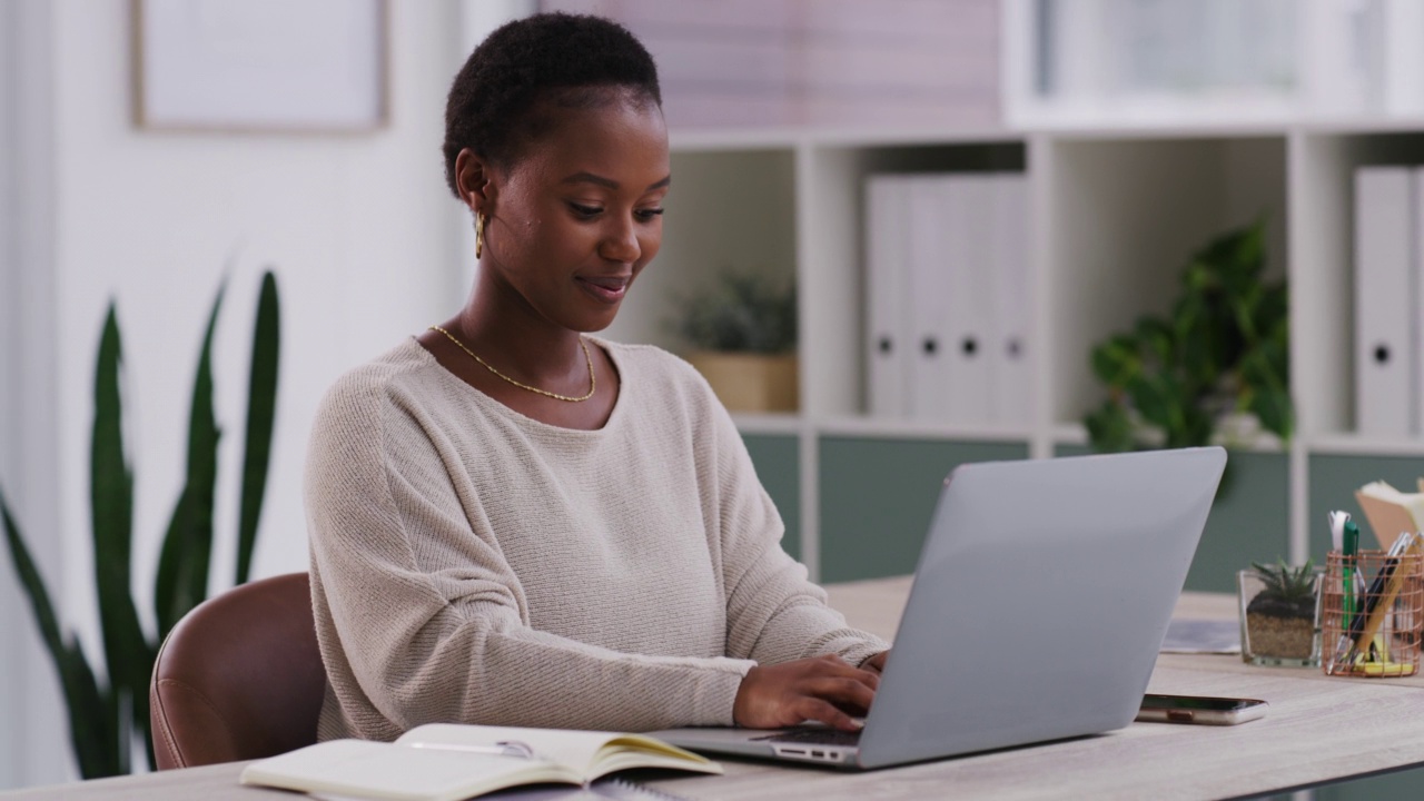 一位非裔美国女商人坐在办公室的办公桌前用笔记本电脑打字。年轻的非洲裔美国女商人在一个现代化的办公室里独自在一台笔记本电脑上工作。视频素材