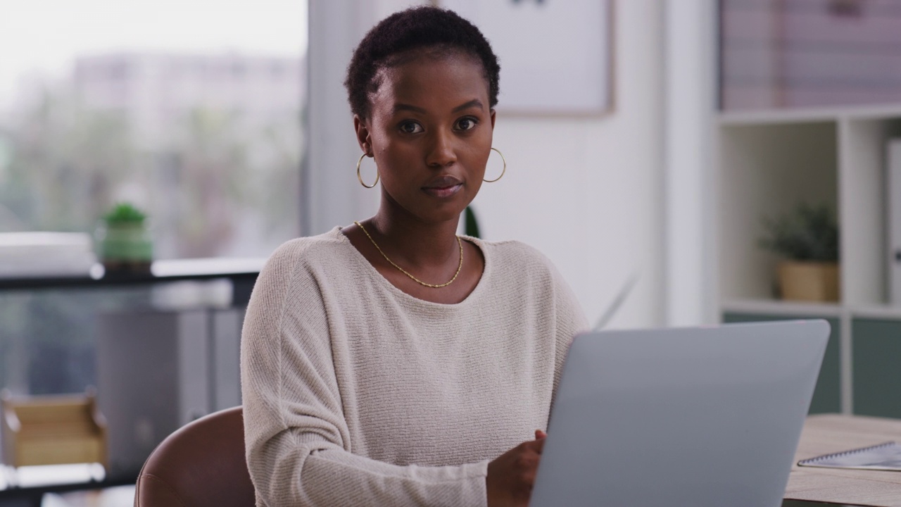 非裔美国女商人，看起来很严肃，在现代办公室里，她坐在书桌前，使用笔记本电脑。一位非裔美国女商人在工作时使用笔记本电脑。视频素材