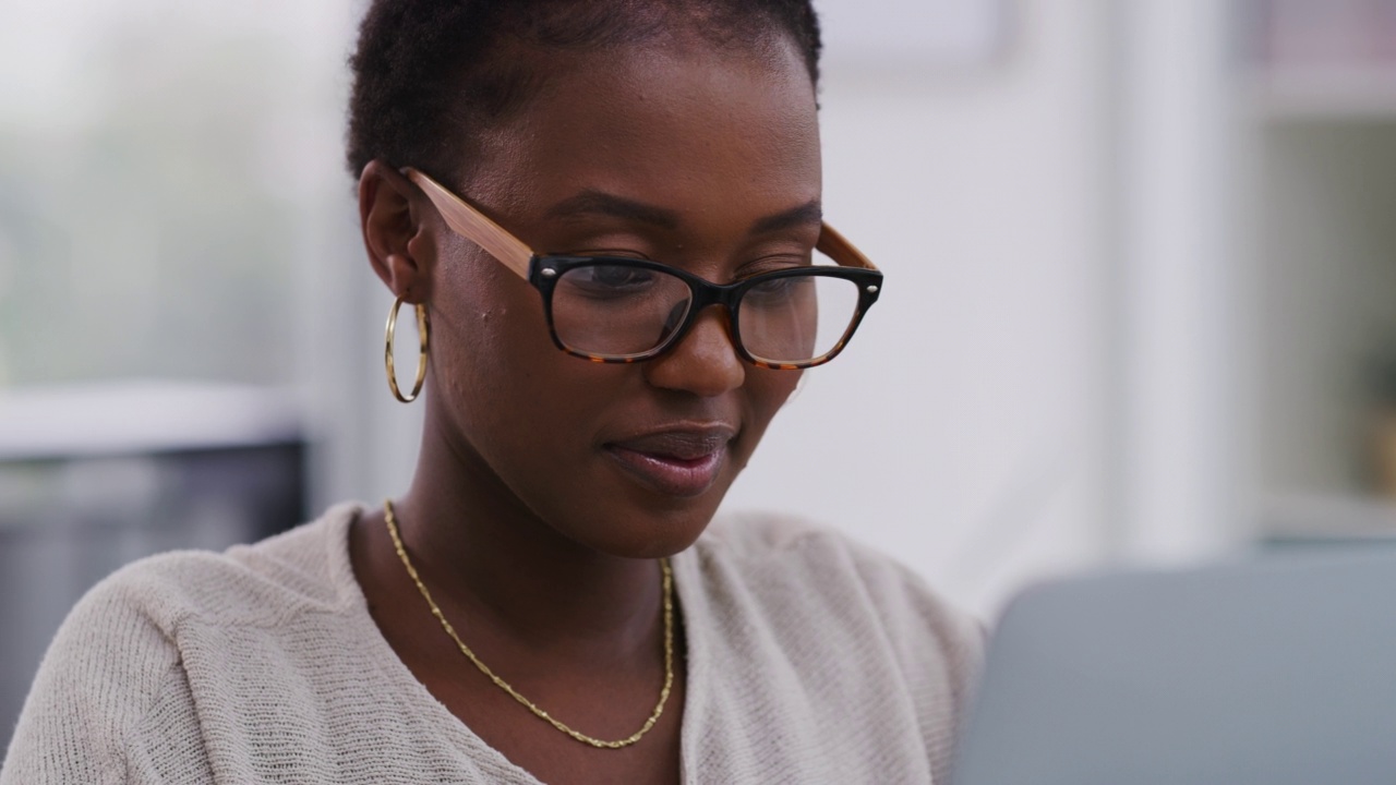 年轻的非洲裔美国女商人微笑着戴眼镜，同时在办公室使用笔记本电脑。一位非裔美国女商人在办公室里用笔记本电脑打字。视频素材