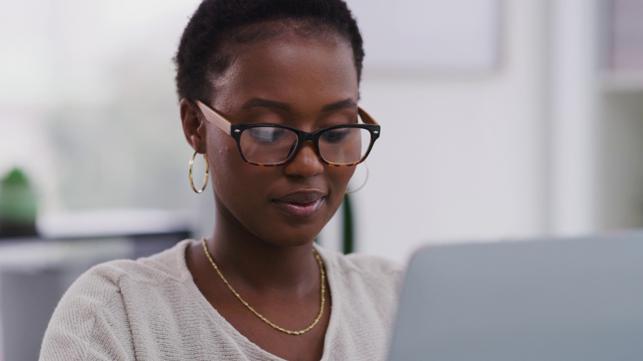 年轻的非洲裔美国女商人戴着眼镜在现代办公室使用笔记本电脑工作。一位非裔美国女商人在工作时微笑着使用笔记本电脑。视频素材