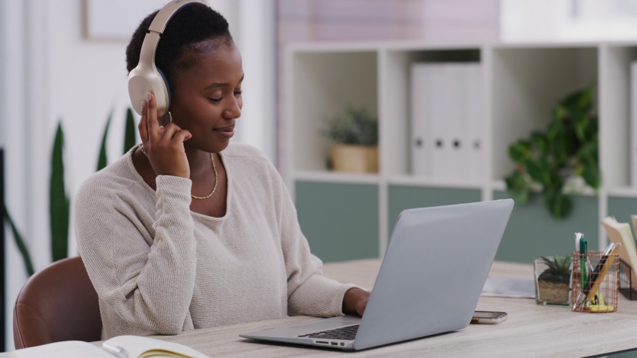 年轻的非洲裔美国女商人在她20多岁的舞蹈享受通过耳机听音乐，同时在现代办公室的数字平板电脑上工作。一位非裔美国女商人在笔记本电脑上打字，听音乐视频素材