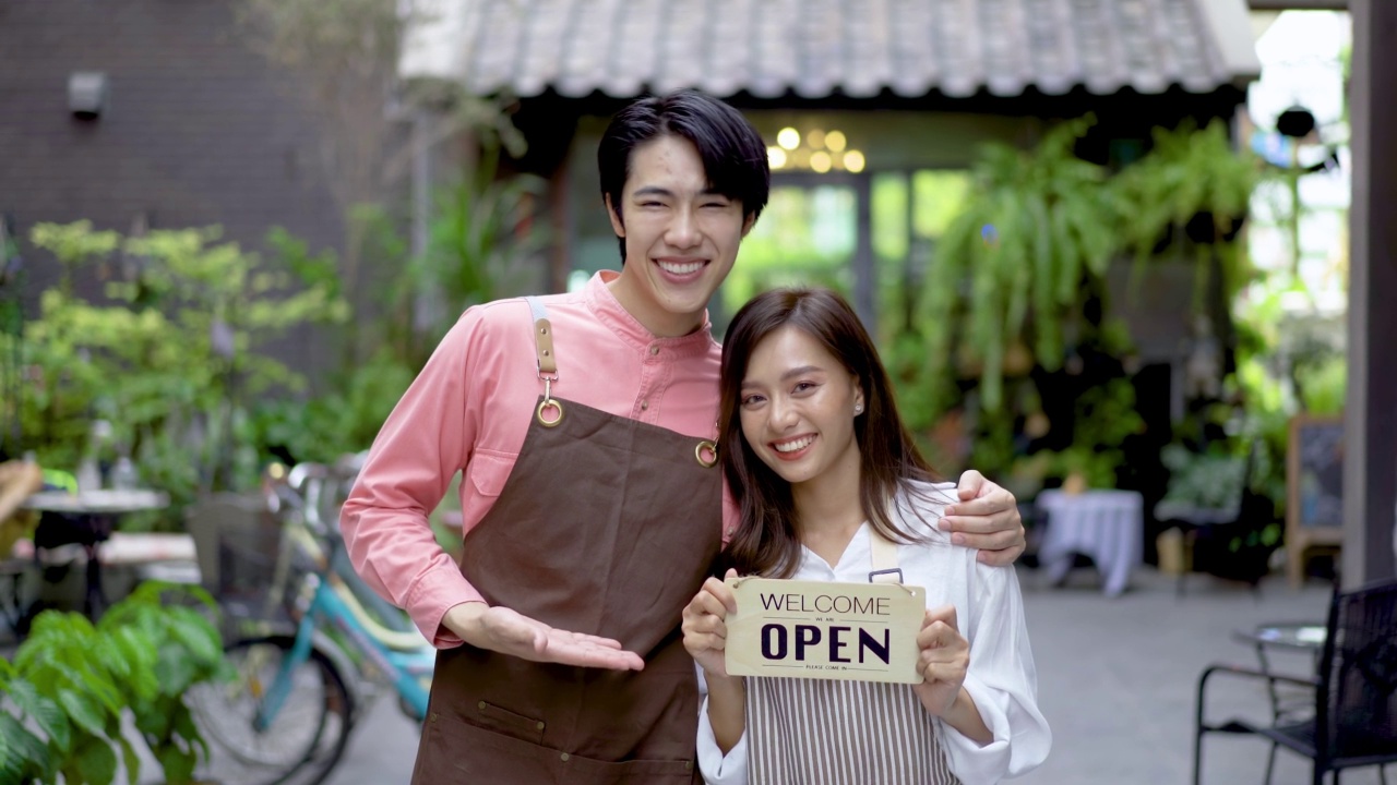 有吸引力的年轻亚洲夫妇很高兴庆祝一家新餐厅的开业，我们在前门的开放标志，微笑着欢迎客户到新的咖啡馆，人和创业视频素材