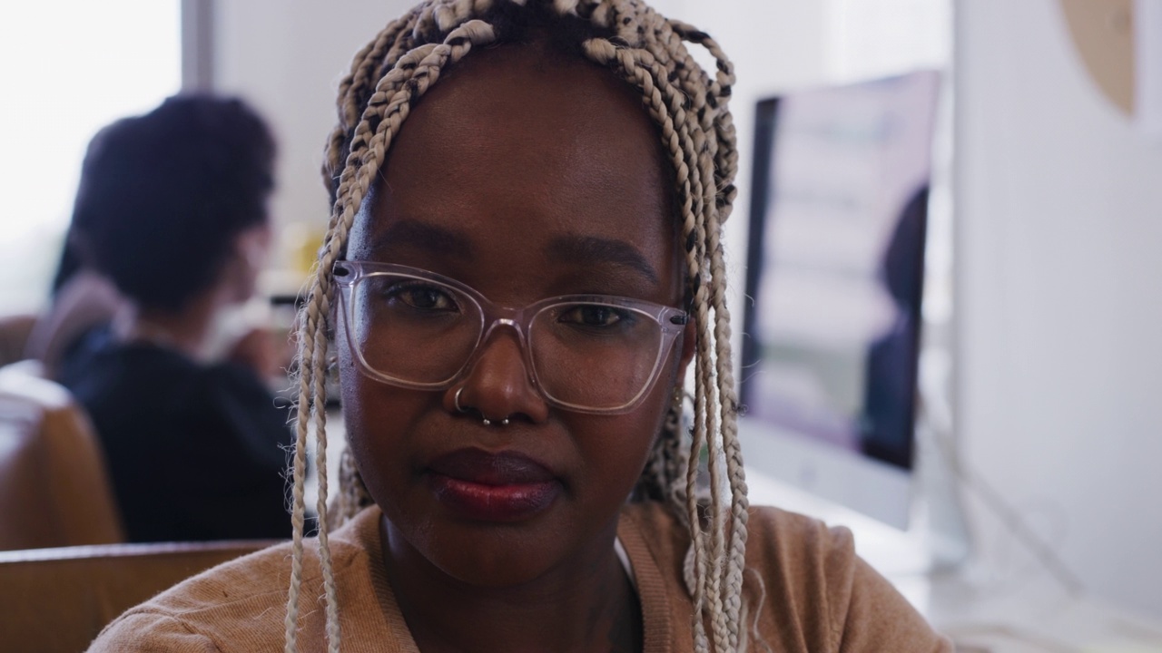 一名黑人妇女戴着眼镜工作的特写肖像，而她的同事在后台的桌面上工作。视频下载