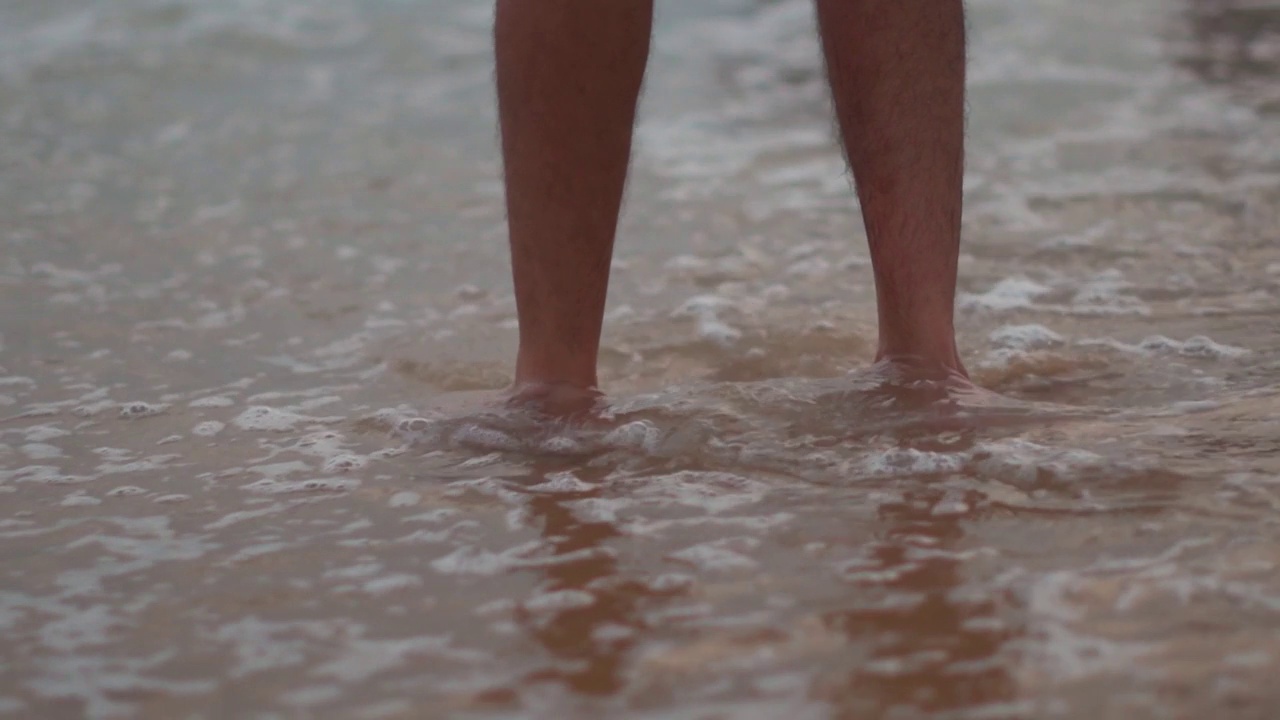 低角度拍摄的腿的一个印度人站在水的海浪的海滩在果阿巴加海滩，印度。一个男人把腿浸在沙滩上的水里。男人正在享受他的暑假。视频素材