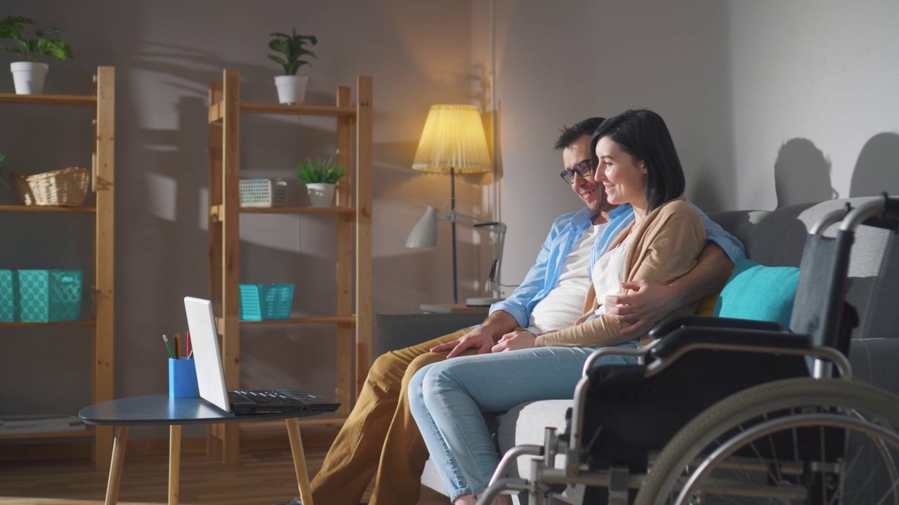 一对夫妇，一个年轻的残疾妇女坐在轮椅上，一起坐在沙发上视频下载