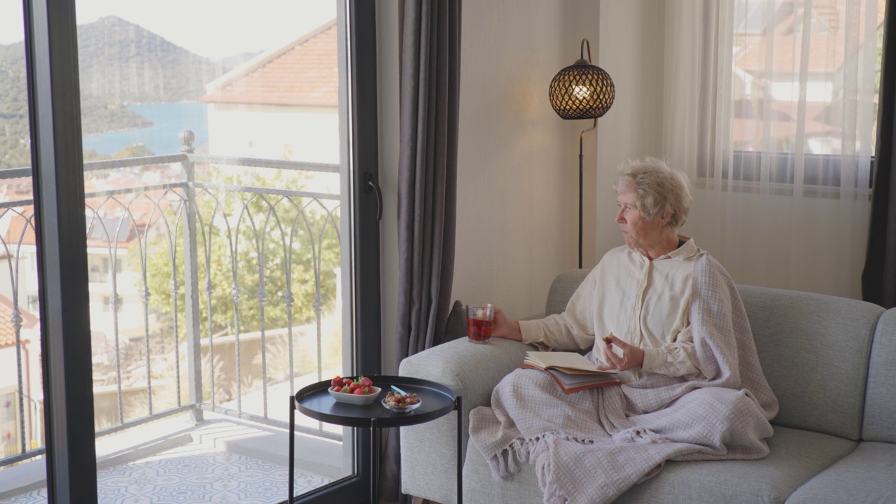 美丽微笑的老女人坐在家里的沙发上喝茶。视频素材
