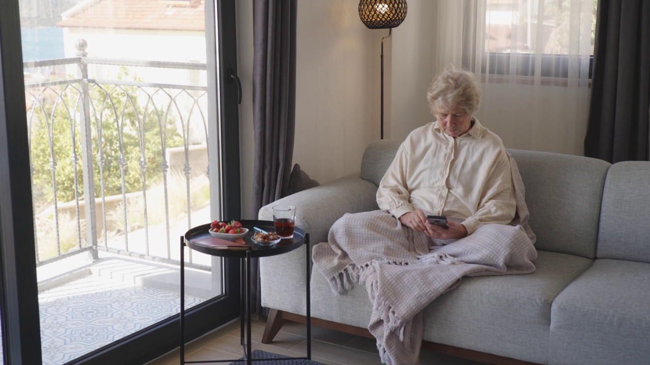 美丽微笑的老女人坐在沙发上，喝茶，用电话在家里。视频素材