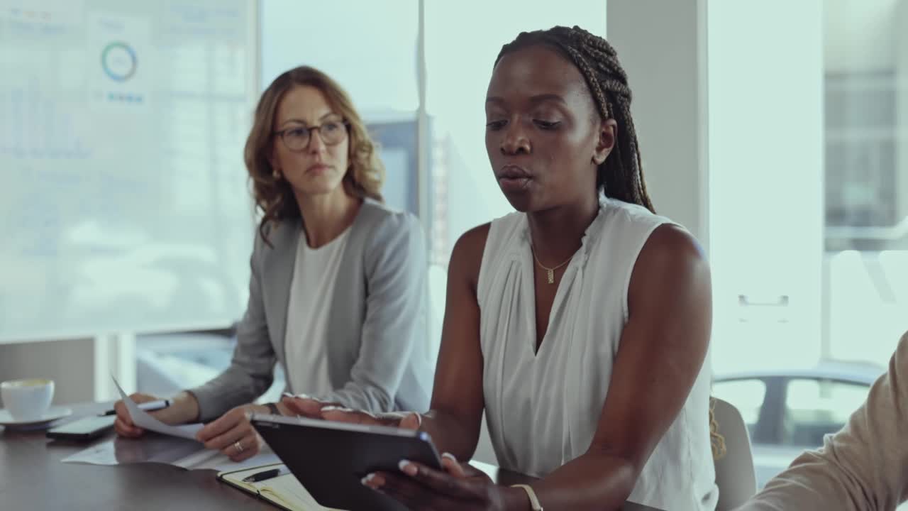 年轻的非洲裔美国女商人，在办公室开会时，说话时看起来很严肃，拿着并向同事展示数字平板电脑屏幕。在工作会议上，不同的同事使用数字平板电脑交谈视频素材