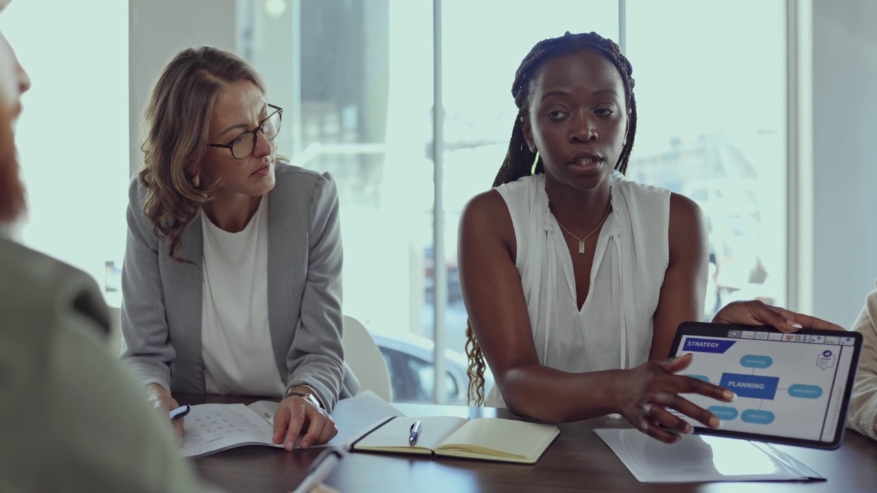年轻的非洲裔美国女商人在工作中与同事在会议室开会时，一边拿着并展示一个数字平板电脑屏幕，一边交谈。商务女性在工作会议上使用数字平板电脑讲话视频素材