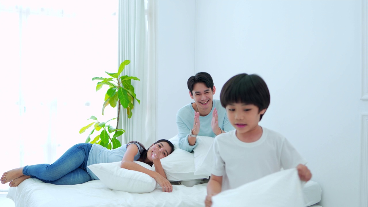 快乐的家庭时光，年轻的亚洲妈妈和爸爸一起看着儿子在卧室玩枕头，开心地笑着。视频素材