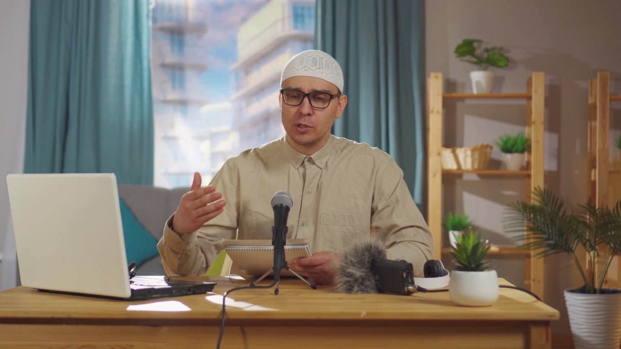 一名戴着眼镜、头戴无边便帽的穆斯林男子坐在客厅的麦克风前录制播客视频下载