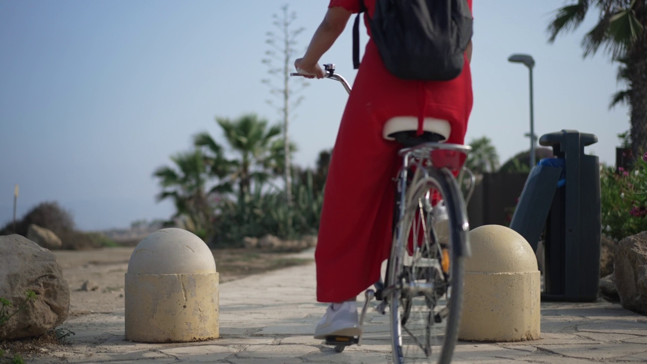 美丽的塞浦路斯风景与年轻女子在红色连衣裙骑自行车通过在慢动作中离开。后视图苗条自信的高加索女性游客骑自行车在夏天的一天。休闲和快乐。视频下载