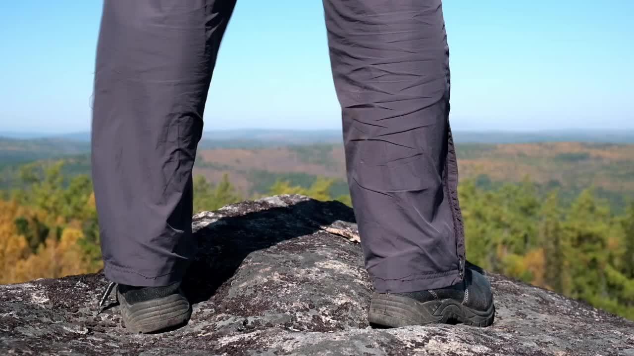 在天空和下面的秋天森林的背景下，一个游客在山顶上徒步旅行的脚的特写。视频素材