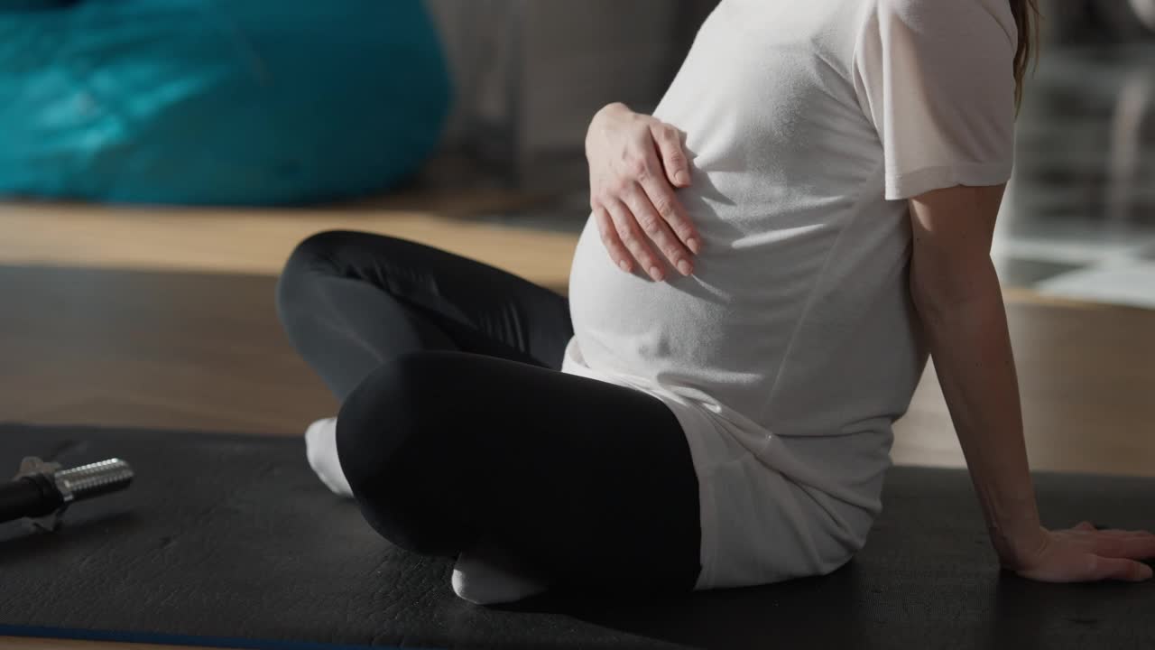 不明身份的怀孕女运动员坐在室内的运动垫子上爱抚腹部。侧视图年轻的高加索妇女休息训练在家里。生活方式和怀孕概念。视频素材