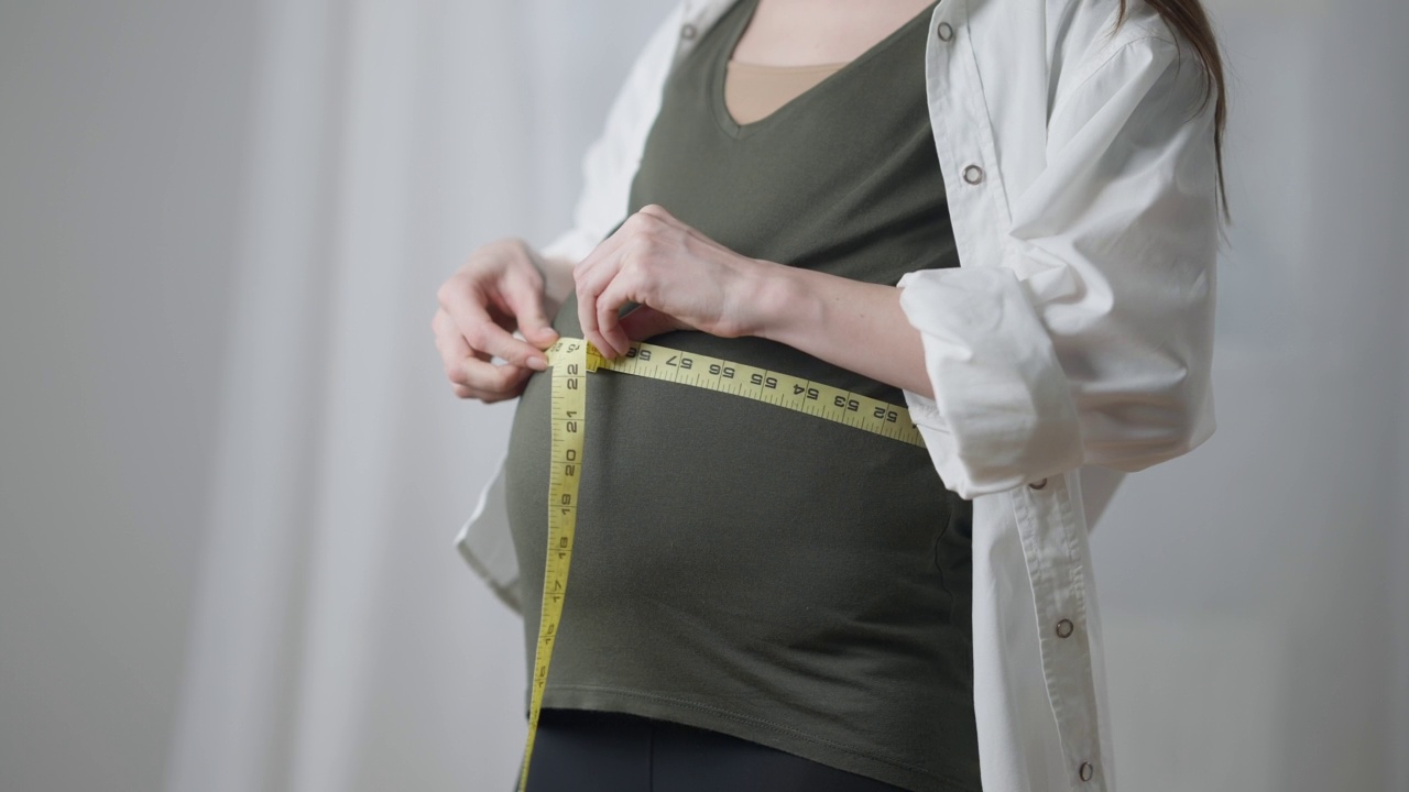 一位不知名的孕妇站在家里量腰围和腹部。白种人年轻苗条的孕妇控制体重和胎儿。保健和怀孕。视频素材