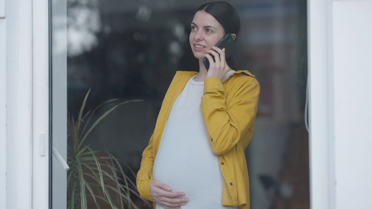 漂亮的孕妇正在讲电话，站在玻璃门向外看。幸福自信的高加索人的肖像，期待享受在家室内休闲。视频素材