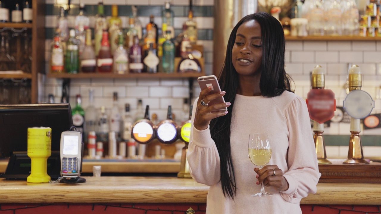 快乐的年轻女子在酒吧里一边用智能手机一边喝白葡萄酒视频下载