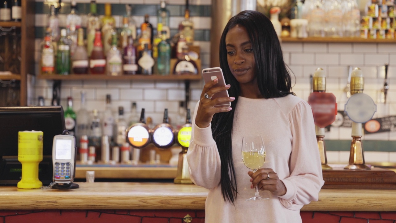 微笑的年轻女子一边用智能手机，一边在酒吧喝着白葡萄酒视频下载
