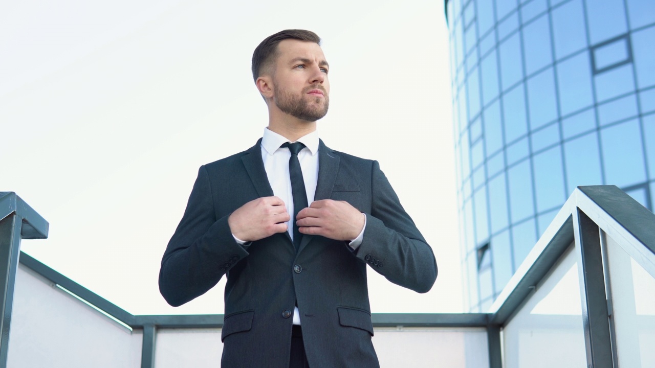 一位穿着得体、满脸胡须的办公室男性高管站在现代公司大楼外，正在调整西装和领带视频下载