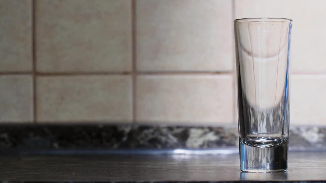 在厨房墙壁的背景下，桌子上有一只玻璃杯，一杯透明液体被倒入其中。伏特加倒入玻璃杯中。一束干净的水倒进玻璃杯里。复制空间的背景视频下载