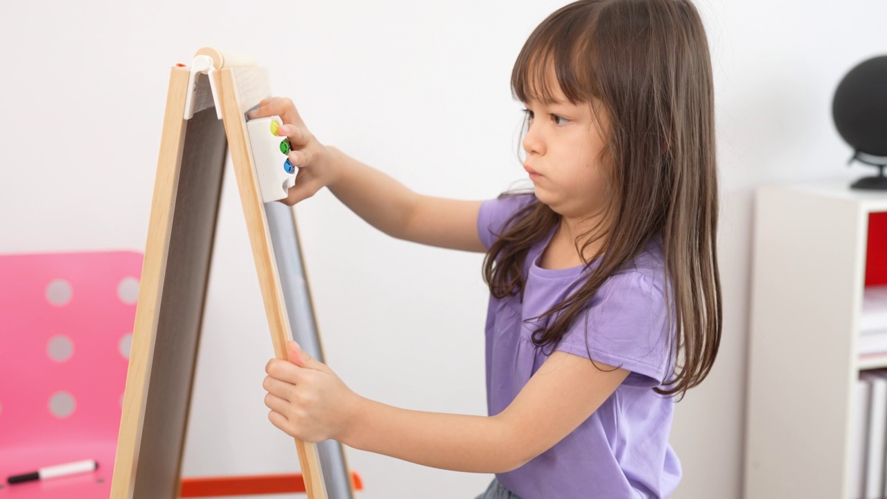 一个可爱的金发小女孩在课堂上用海绵擦黑板的侧面。小女孩在幼儿园学前班上课。初入学校教育，童年教育理念。视频素材