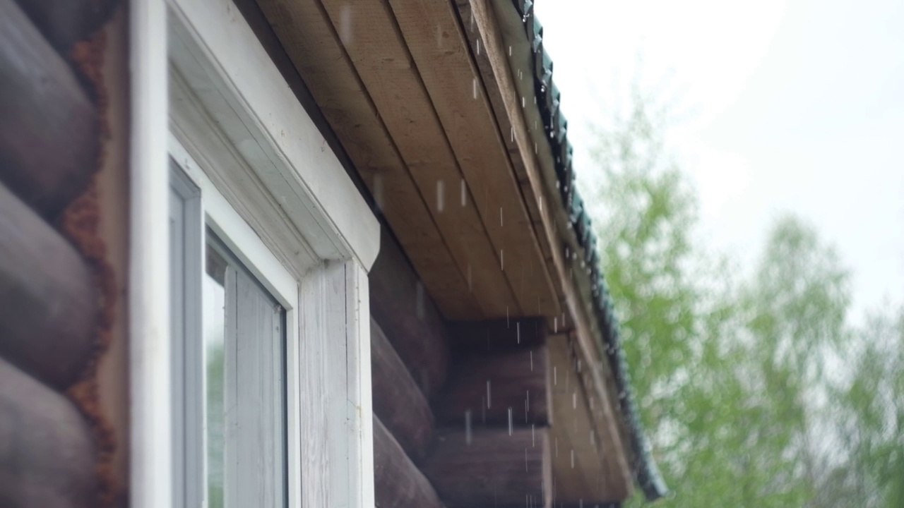 雨水从屋顶流下，以自然的绿色为背景。关闭了。缓慢的运动。夏天阵雨。雨天。棕色的木头房子。雨滴从金属屋顶上快速滴下。侧视图。视频素材
