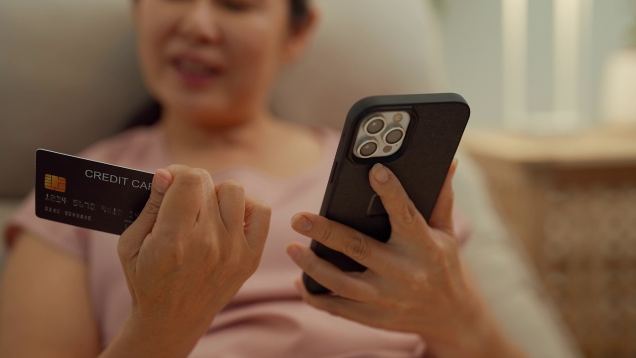 一位年长的亚裔女性站在厨房柜台旁，用智能手机上的移动应用程序(app)订购外卖服务。视频素材