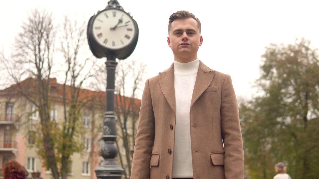 一个英俊时髦的男人的肖像，在街头时钟的背景下，穿着棕色大衣。新收藏的时尚服装视频下载