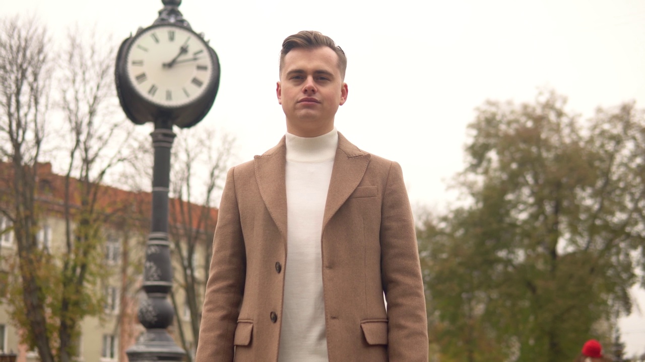 一个英俊时髦的男人的肖像，在街头时钟的背景下，穿着棕色大衣。新收藏的时尚服装视频下载