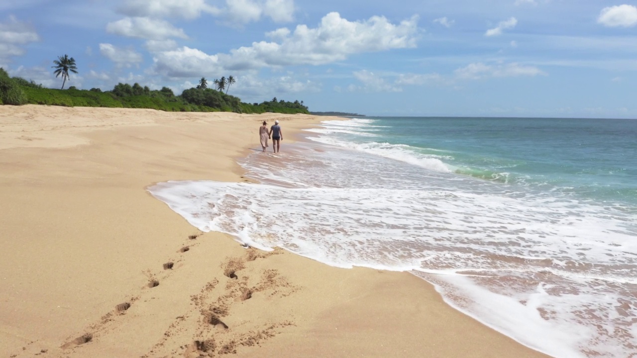幸福的情侣手牵手漫步在印度洋的沙滩上，享受着海浪带来的快乐。斯里兰卡坦加勒附近的4K动态镜头。视频素材