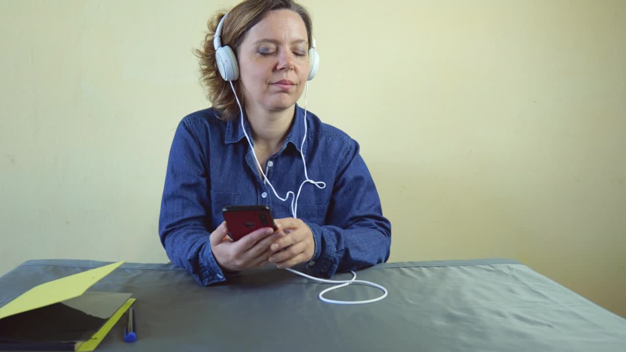 一位戴着白色有线大耳机的40岁妇女正坐在桌旁听音乐。一位成年女性拿着手机视频素材