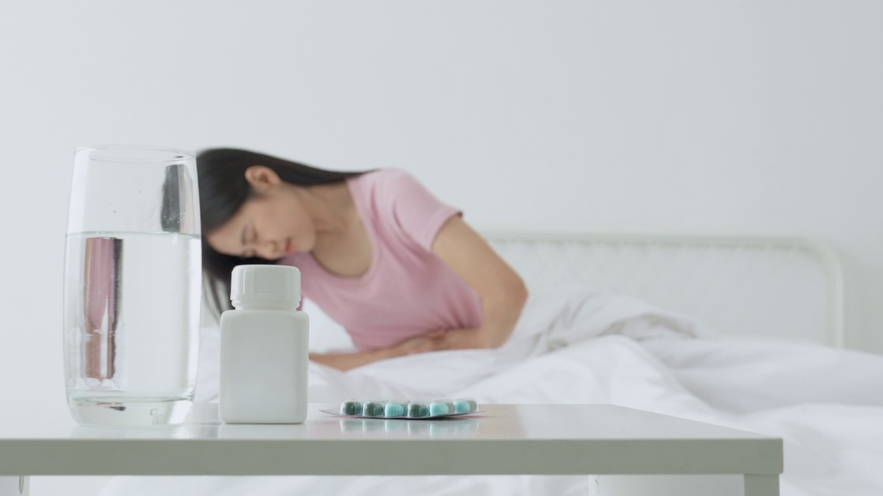 年轻亚洲女性坐在床上腹痛或肠病便秘或消化不良在家卧室，不快乐女性生病和月经和胃痛，医学和健康观念。视频下载