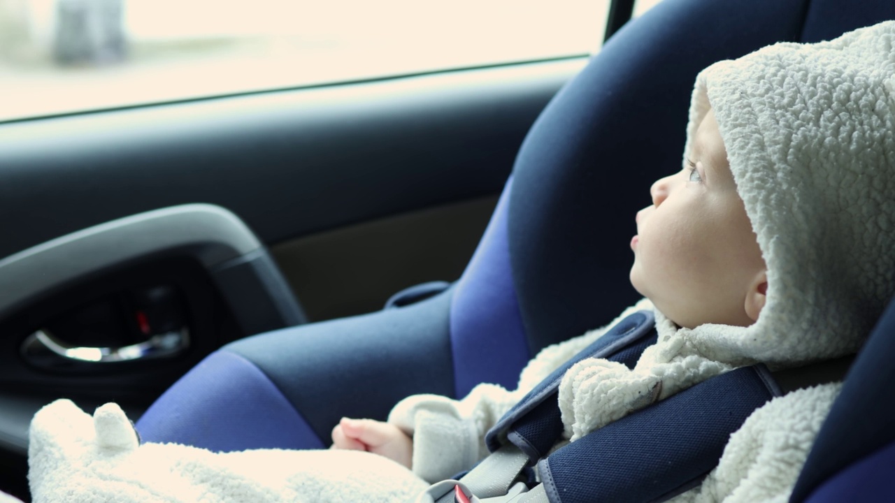 婴儿婴儿在白色的兜帽坐在蓝色的汽车座位上，看着窗户视频素材
