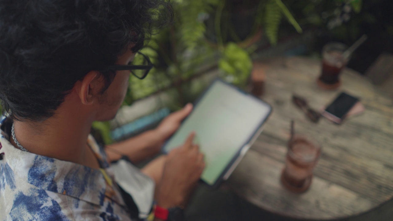 高角度视角:亚洲中年男子戴着休闲布料的眼镜，在一家乡村咖啡馆里一边喝着塑料杯里的冷咖啡，一边用数字平板电脑阅读在线书籍和文字视频素材