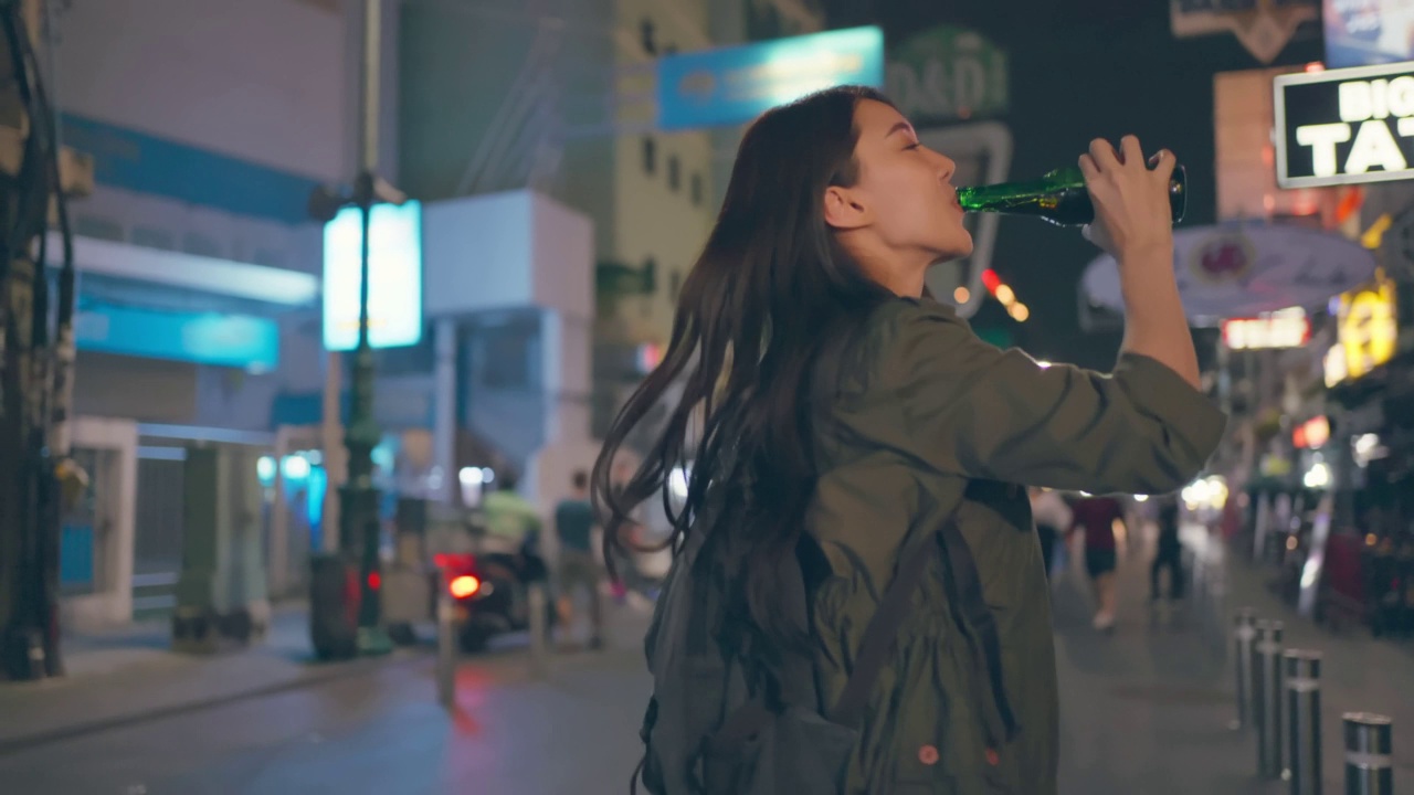 亚洲年轻女性旅行者喝一瓶啤酒在街上走。迷人美丽的女孩旅游旅游在城市，在假期度假和聚会庆祝在黑夜的道路。视频下载