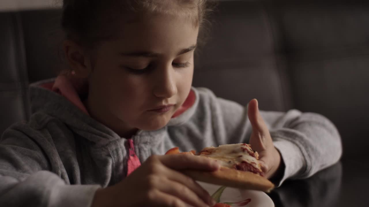 不健康营养快餐。孩子吃午餐的肖像。面对小女孩特写。孩子吃披萨。饥饿的小女孩坐在厨房的餐桌上吃着美味的意大利披萨。视频素材