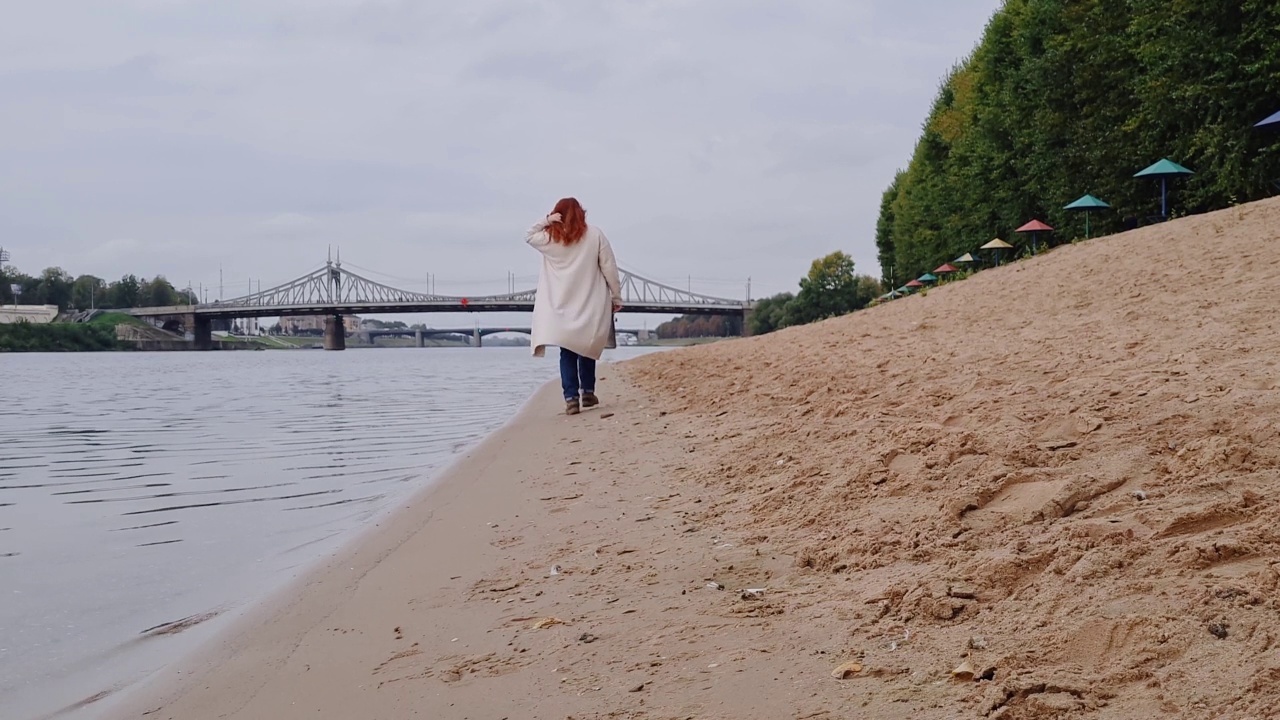 一个年轻的女人正沿着城市的海滩散步。享受秋高气爽的阴天。在大自然中休闲，享受，放松。视频素材