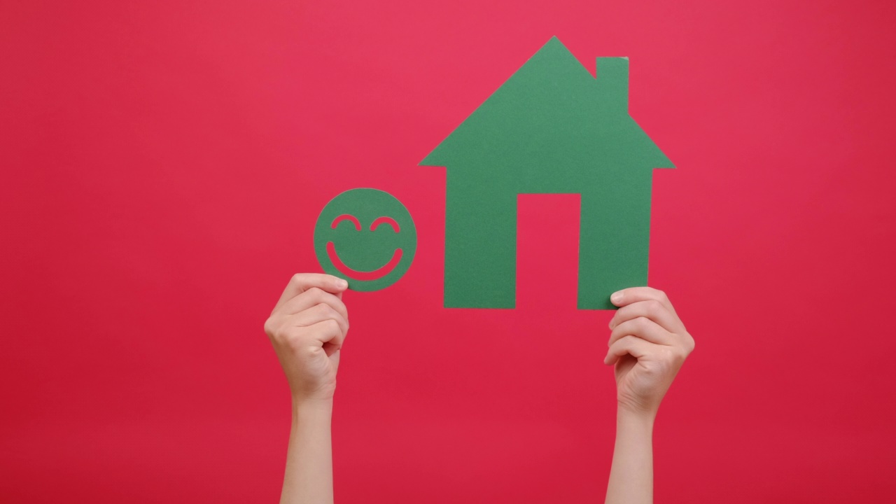 女性双手捧着绿色幸福笑脸纸和房子模型，体验，孤立在红色工作室背景与文案空间的广告。家居、生态、环保的理念视频下载