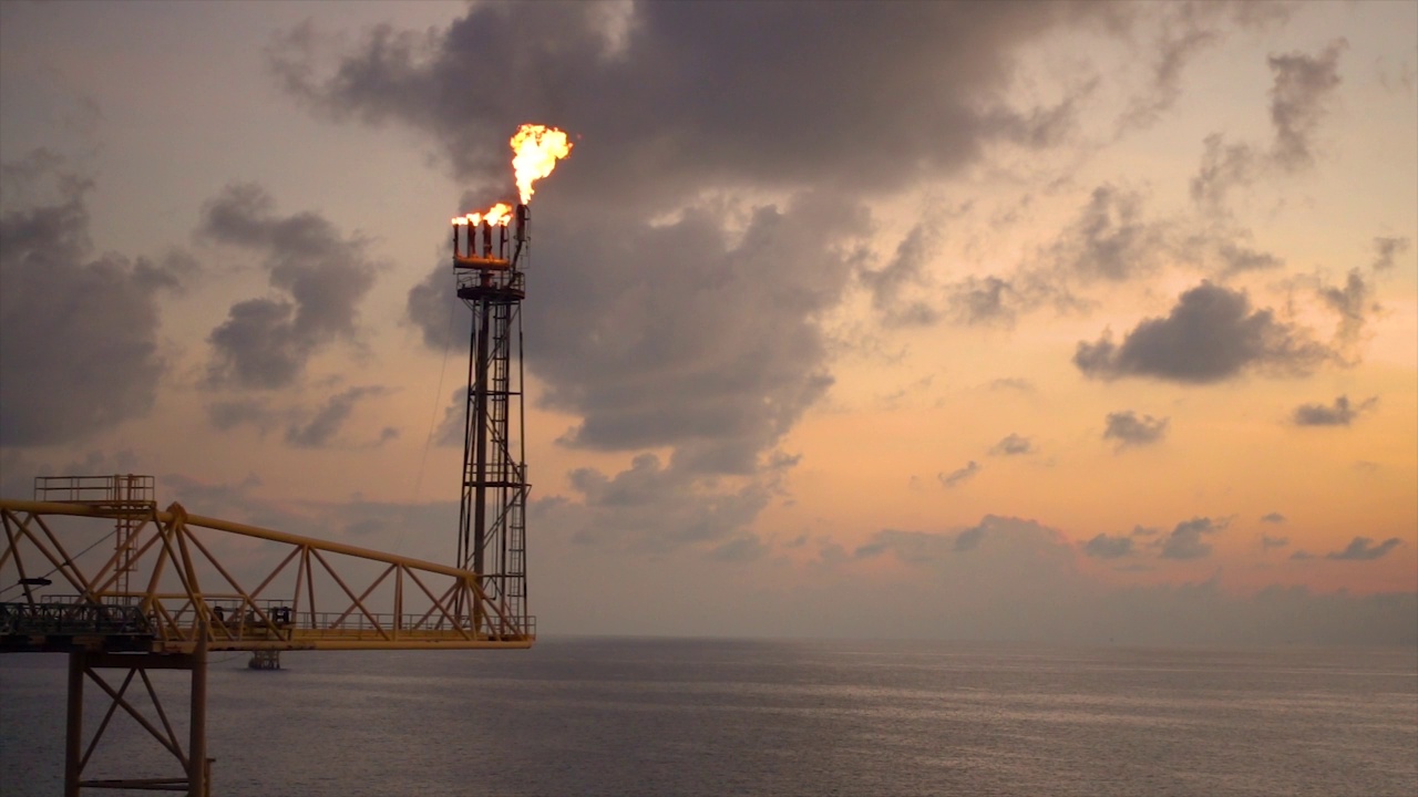在石油和天然气平台底部燃烧的缓慢运动的天然气火炬，背景是夕阳和海洋的石油和天然气业务概念。视频素材
