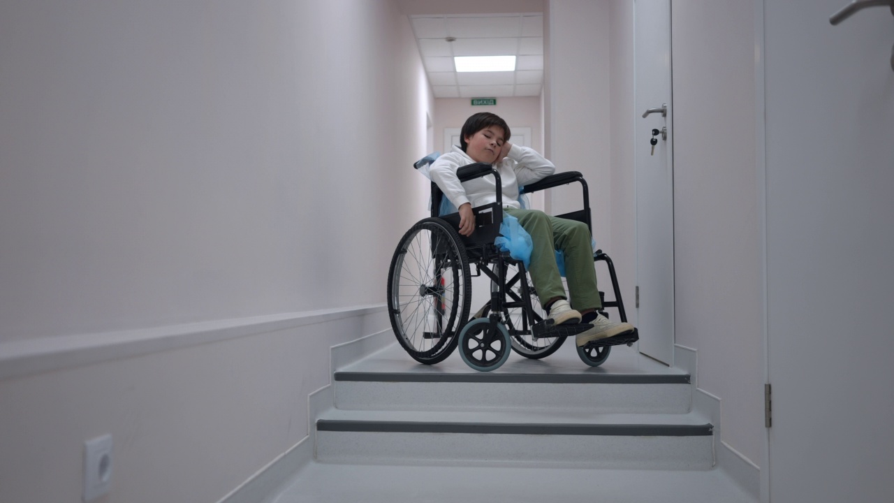 宽镜头肖像悲伤深思的白人男孩坐在轮椅上的楼梯在医院。沮丧可爱的生病孩子思考在医疗诊所走廊。可及性和医学概念。视频素材